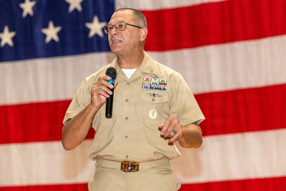 A U.S. Navy senior enlisted or officer personnel is speaking at a formal event while holding a handheld microphone. The individual is wearing a khaki service uniform adorned with multiple ribbons, badges, and insignia. A large American flag serves as the backdrop, suggesting a military ceremony or official address.