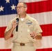 A U.S. Navy senior enlisted or officer personnel is speaking at a formal event while holding a handheld microphone. The individual is wearing a khaki service uniform adorned with multiple ribbons, badges, and insignia. A large American flag serves as the backdrop, suggesting a military ceremony or official address.