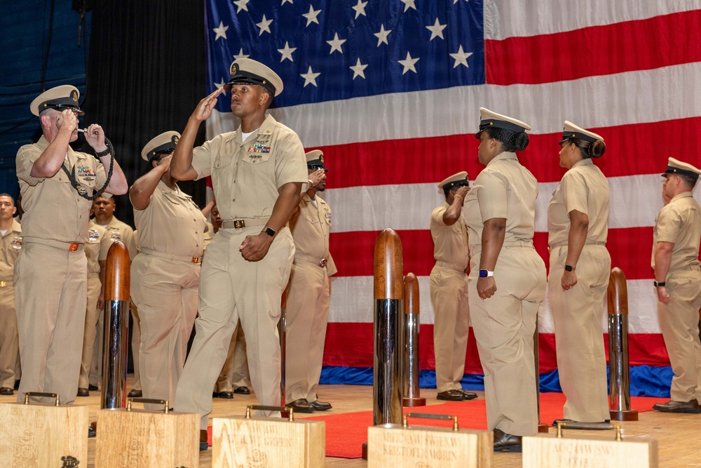 A formal U.S. Navy ceremony is taking place on a stage in front of a large American flag backdrop. Multiple Navy personnel in khaki service uniforms are rendering salutes, suggesting a military ceremony such as a promotion, retirement, or chief petty officer pinning event. Decorative wooden and metal posts with plaques are arranged in the foreground of the stage.