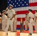 A formal U.S. Navy ceremony is taking place on a stage in front of a large American flag backdrop. Multiple Navy personnel in khaki service uniforms are rendering salutes, suggesting a military ceremony such as a promotion, retirement, or chief petty officer pinning event. Decorative wooden and metal posts with plaques are arranged in the foreground of the stage.