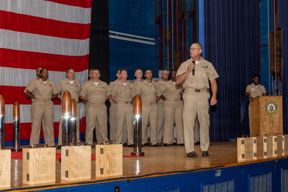 A U.S. Navy ceremony taking place on a stage, with a senior officer speaking into a microphone while a group of sailors in tan/khaki uniforms stand at attention behind him. Several wooden award plaques or trophies with metallic bullet-shaped tops are displayed in the foreground on the stage. A large American flag serves as a backdrop on the left side of the stage.