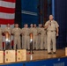 A U.S. Navy ceremony taking place on a stage, with a senior officer speaking into a microphone while a group of sailors in tan/khaki uniforms stand at attention behind him. Several wooden award plaques or trophies with metallic bullet-shaped tops are displayed in the foreground on the stage. A large American flag serves as a backdrop on the left side of the stage.