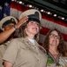 A U.S. Navy ceremony is taking place in front of a large American flag. A female Navy chief petty officer in khaki uniform is saluting while adjusting her combination cover (hat), with ribbons/medals visible on her chest. A civilian woman and another uniformed Navy sailor are visible beside and behind her.