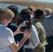 A crowd of people, including at least one individual in military camouflage uniform, are gathered outdoors at what appears to be a public event or demonstration. Several attendees are holding up smartphones to photograph or record something out of frame. The setting appears to be a sunny day with mountains and a structure visible in the background.