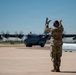 A military service member in OCP (Operational Camouflage Pattern) uniform is marshaling a large military turboprop aircraft on a flight line. The individual is using red marshaling wands with both arms raised, guiding the aircraft toward its parking position. The scene takes place on a concrete tarmac under clear blue skies.