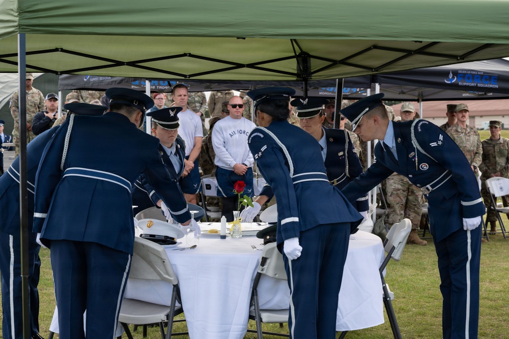 Military personnel in formal dress uniforms are conducting a POW/MIA Missing Man Table ceremony outdoors under a green canopy tent. Several Air Force members in dark blue ceremonial uniforms with white gloves are arranging items on a small round table covered with a white tablecloth. A crowd of uniformed military observers watches from the background.