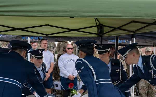 Military personnel in formal dress uniforms are conducting a POW/MIA Missing Man Table ceremony outdoors under a green canopy tent. Several Air Force members in dark blue ceremonial uniforms with white gloves are arranging items on a small round table covered with a white tablecloth. A crowd of uniformed military observers watches from the background.