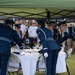 Military personnel in formal dress uniforms are conducting a POW/MIA Missing Man Table ceremony outdoors under a green canopy tent. Several Air Force members in dark blue ceremonial uniforms with white gloves are arranging items on a small round table covered with a white tablecloth. A crowd of uniformed military observers watches from the background.