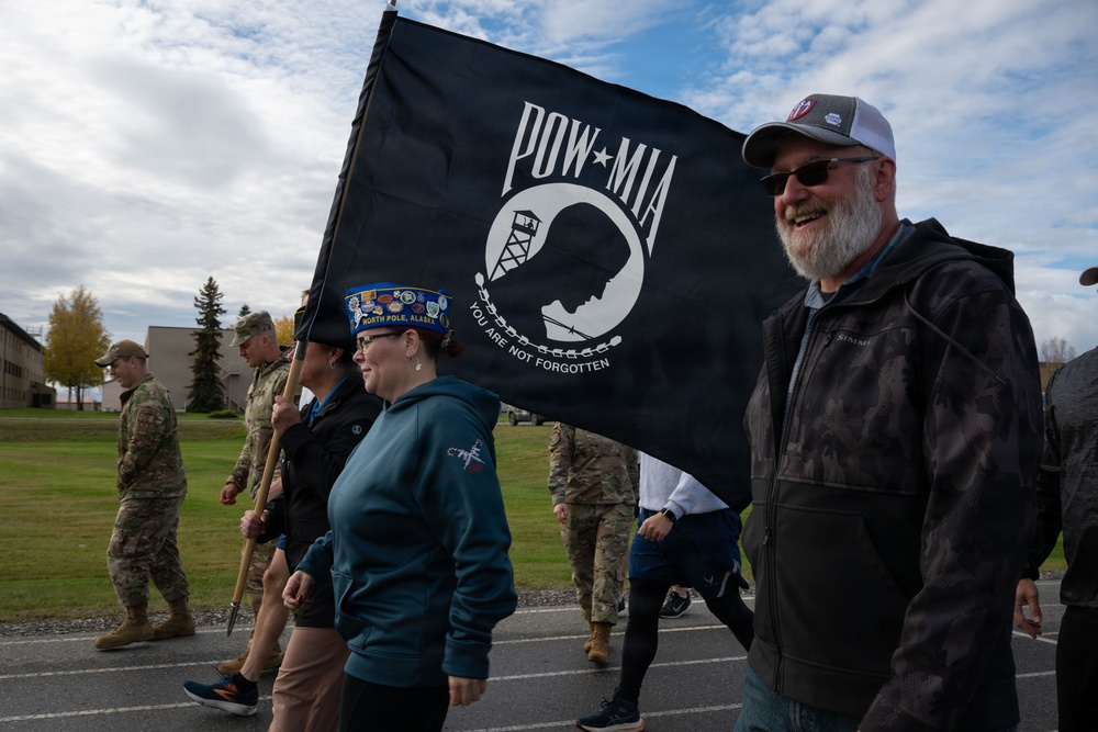 A group of civilians and military personnel participate in a commemorative march or walk, carrying a black POW-MIA flag reading 'You Are Not Forgotten.' The event appears to take place on a military installation, likely in Alaska based on the visible 'North Pole, Alaska' text on a participant's hat. The atmosphere is solemn yet communal, honoring prisoners of war and those missing in action.