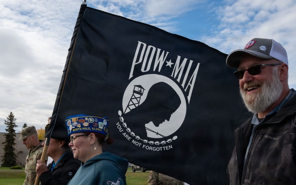 A group of civilians and military personnel participate in a commemorative march or walk, carrying a black POW-MIA flag reading 'You Are Not Forgotten.' The event appears to take place on a military installation, likely in Alaska based on the visible 'North Pole, Alaska' text on a participant's hat. The atmosphere is solemn yet communal, honoring prisoners of war and those missing in action.