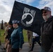 A group of civilians and military personnel participate in a commemorative march or walk, carrying a black POW-MIA flag reading 'You Are Not Forgotten.' The event appears to take place on a military installation, likely in Alaska based on the visible 'North Pole, Alaska' text on a participant's hat. The atmosphere is solemn yet communal, honoring prisoners of war and those missing in action.