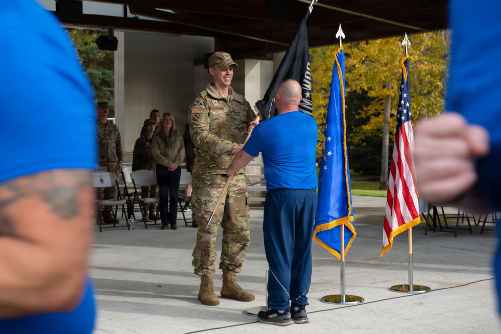 A military ceremony is taking place outdoors under a covered pavilion, where a uniformed soldier in OCP (Operational Camouflage Pattern) is receiving or exchanging a dark flag with a civilian or off-duty service member in a blue shirt. The scene includes multiple flags including the United States flag and what appears to be an Air Force flag, suggesting this may be a change of command or retirement ceremony. Spectators in military and civilian attire are visible in the background.