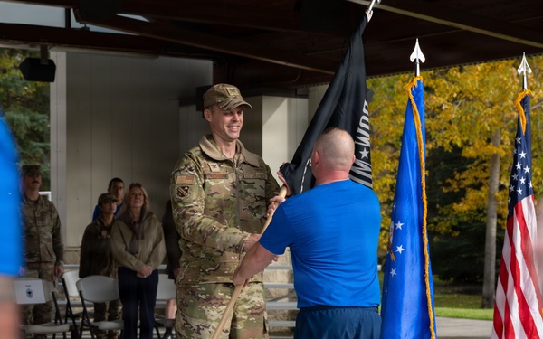 A military ceremony is taking place outdoors under a covered pavilion, where a uniformed soldier in OCP (Operational Camouflage Pattern) is receiving or exchanging a dark flag with a civilian or off-duty service member in a blue shirt. The scene includes multiple flags including the United States flag and what appears to be an Air Force flag, suggesting this may be a change of command or retirement ceremony. Spectators in military and civilian attire are visible in the background.