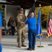 A military ceremony is taking place outdoors under a covered pavilion, where a uniformed soldier in OCP (Operational Camouflage Pattern) is receiving or exchanging a dark flag with a civilian or off-duty service member in a blue shirt. The scene includes multiple flags including the United States flag and what appears to be an Air Force flag, suggesting this may be a change of command or retirement ceremony. Spectators in military and civilian attire are visible in the background.