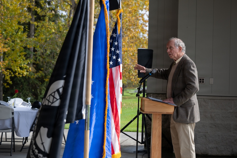 An elderly man in a brown tweed blazer speaks at a wooden podium during what appears to be an outdoor ceremonial event. Multiple flags are displayed prominently, including the United States flag, a blue flag with gold fringe, and a black POW/MIA flag. The setting includes autumn foliage in the background and a white-clothed table visible to the left.