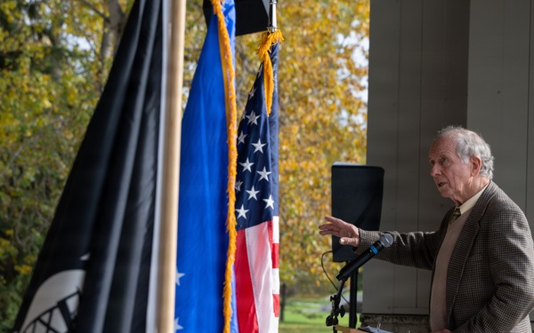 An elderly man in a brown tweed blazer speaks at a wooden podium during what appears to be an outdoor ceremonial event. Multiple flags are displayed prominently, including the United States flag, a blue flag with gold fringe, and a black POW/MIA flag. The setting includes autumn foliage in the background and a white-clothed table visible to the left.