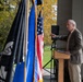 An elderly man in a brown tweed blazer speaks at a wooden podium during what appears to be an outdoor ceremonial event. Multiple flags are displayed prominently, including the United States flag, a blue flag with gold fringe, and a black POW/MIA flag. The setting includes autumn foliage in the background and a white-clothed table visible to the left.