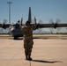 A military ground crew member in camouflage uniform is marshalling a C-130 Hercules turboprop military transport aircraft on a flight line. The airman, facing away from the camera, holds red marshalling wands with both arms raised above their head. The aircraft's four propellers are visibly spinning, indicating the engines are running.
