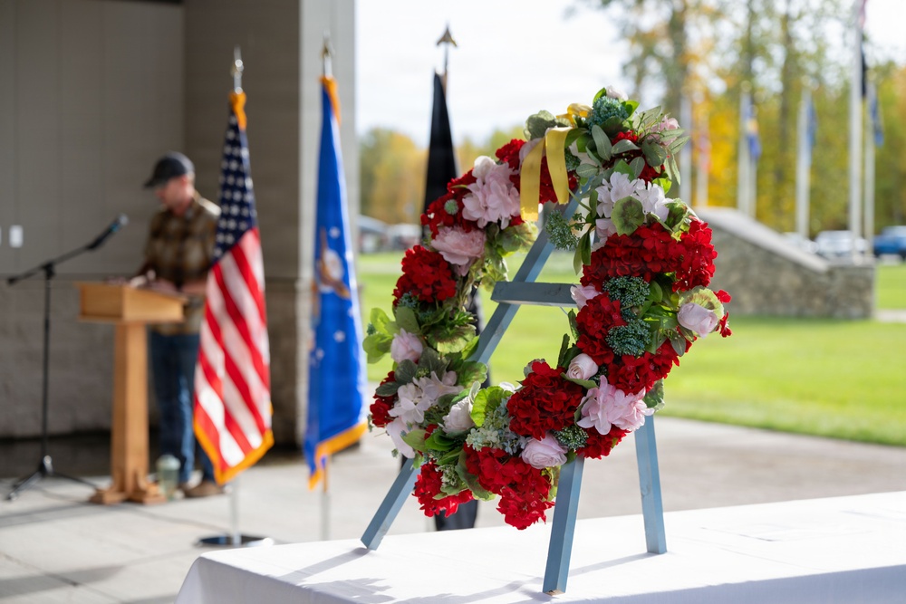 A formal memorial or veterans ceremony is taking place outdoors under a covered pavilion. A floral wreath composed of red, pink, and white flowers with a yellow ribbon is displayed prominently in the foreground on a blue easel stand. A speaker addresses the gathering from a podium in the background, flanked by the American flag and what appears to be a military branch flag.