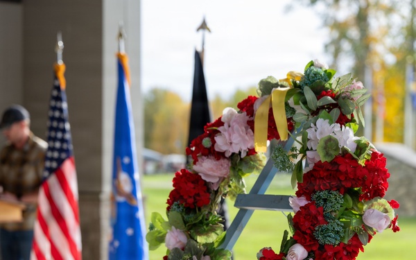 A formal memorial or veterans ceremony is taking place outdoors under a covered pavilion. A floral wreath composed of red, pink, and white flowers with a yellow ribbon is displayed prominently in the foreground on a blue easel stand. A speaker addresses the gathering from a podium in the background, flanked by the American flag and what appears to be a military branch flag.