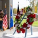 A formal memorial or veterans ceremony is taking place outdoors under a covered pavilion. A floral wreath composed of red, pink, and white flowers with a yellow ribbon is displayed prominently in the foreground on a blue easel stand. A speaker addresses the gathering from a podium in the background, flanked by the American flag and what appears to be a military branch flag.