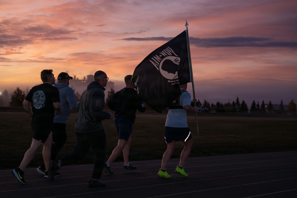 A group of people are running on a track at dawn or dusk while one participant carries a POW/MIA flag. The scene appears to be a memorial or commemorative run, likely held on a military installation given the flag and setting. The dramatic pink and purple sky creates a striking backdrop for what appears to be a solemn, purposeful event.