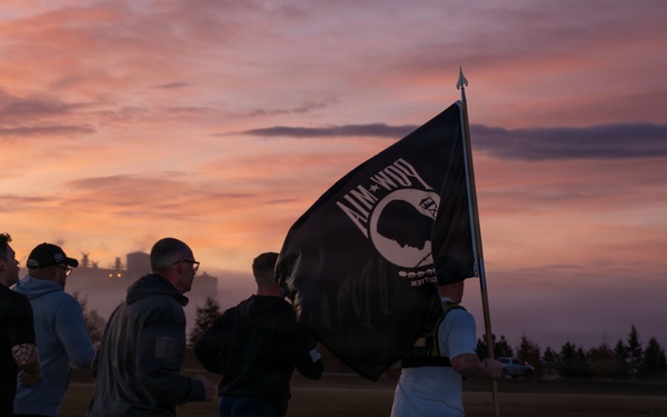 A group of people are running on a track at dawn or dusk while one participant carries a POW/MIA flag. The scene appears to be a memorial or commemorative run, likely held on a military installation given the flag and setting. The dramatic pink and purple sky creates a striking backdrop for what appears to be a solemn, purposeful event.