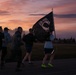 A group of people are running on a track at dawn or dusk while one participant carries a POW/MIA flag. The scene appears to be a memorial or commemorative run, likely held on a military installation given the flag and setting. The dramatic pink and purple sky creates a striking backdrop for what appears to be a solemn, purposeful event.