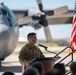 A U.S. military service member in OCP (Operational Camouflage Pattern) uniform is delivering a speech at a podium on a flight line, with a large turboprop military aircraft (likely a C-130 Hercules) visible in the background. An American flag is displayed to the right of the speaker, suggesting a formal military ceremony or official event. An audience is partially visible in the foreground.