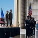 A formal U.S. Air Force military ceremony is taking place inside or at the entrance of a large aircraft hangar. A color guard in dress blue uniforms is marching with the U.S. flag and an Air Force flag, while two officers in flight suits stand at attention on a raised stage. The setting and elements suggest a change of command or similar official military ceremony.