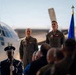 A formal U.S. Air Force military ceremony taking place on a flight line or tarmac, with two officers in olive green flight suits standing at attention before an audience. A uniformed honor guard member is visible in the foreground holding a ceremonial rifle, and a blue military unit flag is displayed to the right. A large military transport aircraft, likely a C-130, is visible in the background.