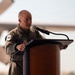 A bald military officer in an olive/sage green flight suit is speaking at a wooden podium equipped with a microphone. He appears to be delivering a speech or address at an outdoor ceremony, with what appears to be a military aircraft visible in the blurred background. Multiple unit patches are visible on his flight suit.