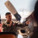 A U.S. military officer in a flight suit is delivering a speech at a podium equipped with a microphone, pointing toward the audience. The setting appears to be an aircraft hangar or flight line, with a large propeller-driven aircraft visible in the background. An American flag with a gold fringe stands to the speaker's left.