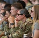 A group of military personnel in OCP (Operational Camouflage Pattern) uniforms are seated in what appears to be bleachers or stadium-style seating, attentively watching an event or presentation. Several civilians are also present among the group. The individuals appear engaged and focused, with contemplative expressions and hands near their faces.