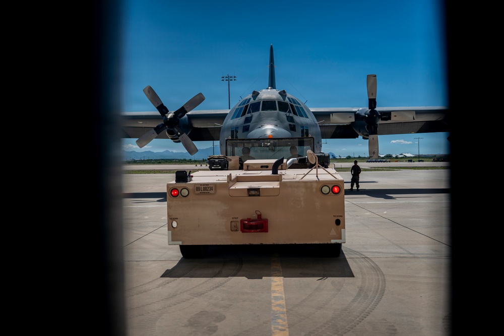 A military transport aircraft, identifiable as a C-130 Hercules, is being towed on an airfield tarmac by a ground support tug vehicle. The image is captured through a framed opening, likely a door or hatch, giving a bordered perspective. A ground crew member is visible standing near the aircraft on the right side.