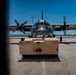 A military transport aircraft, identifiable as a C-130 Hercules, is being towed on an airfield tarmac by a ground support tug vehicle. The image is captured through a framed opening, likely a door or hatch, giving a bordered perspective. A ground crew member is visible standing near the aircraft on the right side.