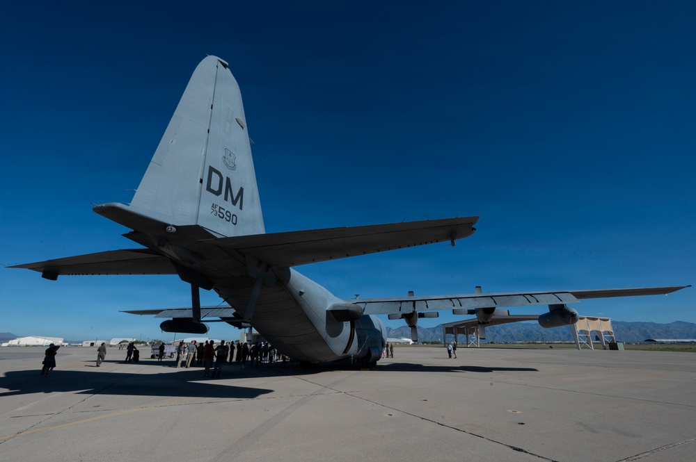 A large military transport aircraft, identifiable as a C-130 Hercules, is parked on a tarmac at what appears to be Davis-Monthan Air Force Base (indicated by the 'DM' tail code and serial number 590). The aircraft is photographed from a low rear angle, emphasizing the tail section and wings. A group of civilians and/or military personnel are gathered near the rear of the aircraft, suggesting a public event, tour, or static display.
