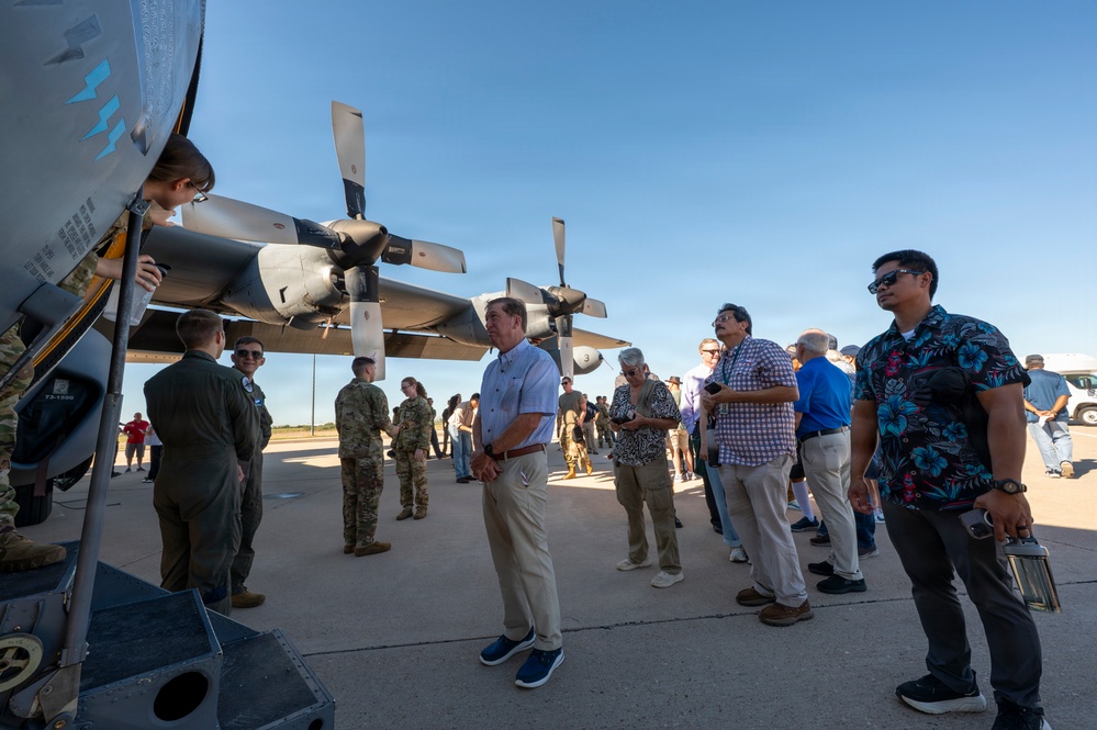 A group of civilians and military personnel are gathered on an airfield tarmac for what appears to be an aircraft static display or base tour event. They are examining the underside and engines of a large military turboprop aircraft, likely a C-130 Hercules, with propeller assemblies clearly visible. The mix of uniformed service members and casually dressed civilians suggests an organized community outreach or civic leader orientation event.