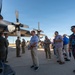 A group of civilians and military personnel are gathered on an airfield tarmac for what appears to be an aircraft static display or base tour event. They are examining the underside and engines of a large military turboprop aircraft, likely a C-130 Hercules, with propeller assemblies clearly visible. The mix of uniformed service members and casually dressed civilians suggests an organized community outreach or civic leader orientation event.