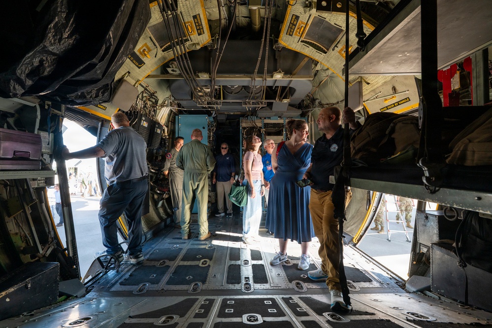 A group of civilians and military personnel are touring the interior cargo bay of a large military transport aircraft, likely a C-130 Hercules. The visitors appear to be on an organized tour or open house event, examining the aircraft's interior systems and equipment. Both uniformed military members and civilian guests wearing ID badges are present.