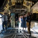 A group of civilians and military personnel are touring the interior cargo bay of a large military transport aircraft, likely a C-130 Hercules. The visitors appear to be on an organized tour or open house event, examining the aircraft's interior systems and equipment. Both uniformed military members and civilian guests wearing ID badges are present.