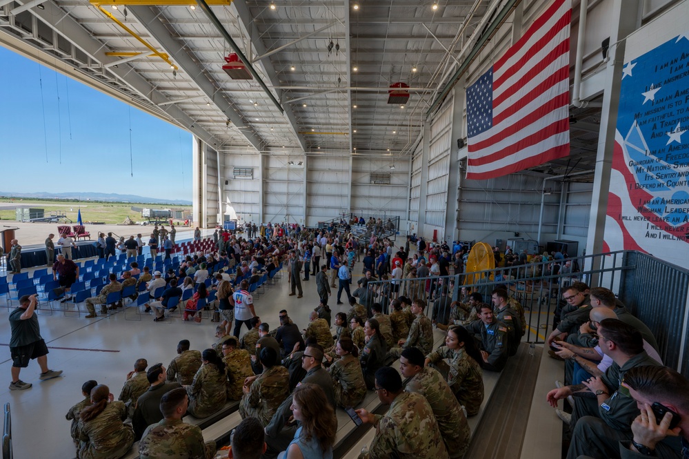 A large military ceremony or gathering is taking place inside an aircraft hangar, with hundreds of uniformed military personnel and civilians in attendance. A large American flag is prominently displayed on the right wall alongside what appears to be the Airman's Creed text. The hangar doors are open, revealing an airfield and blue sky in the background.