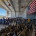 A large military ceremony or gathering is taking place inside an aircraft hangar, with hundreds of uniformed military personnel and civilians in attendance. A large American flag is prominently displayed on the right wall alongside what appears to be the Airman's Creed text. The hangar doors are open, revealing an airfield and blue sky in the background.