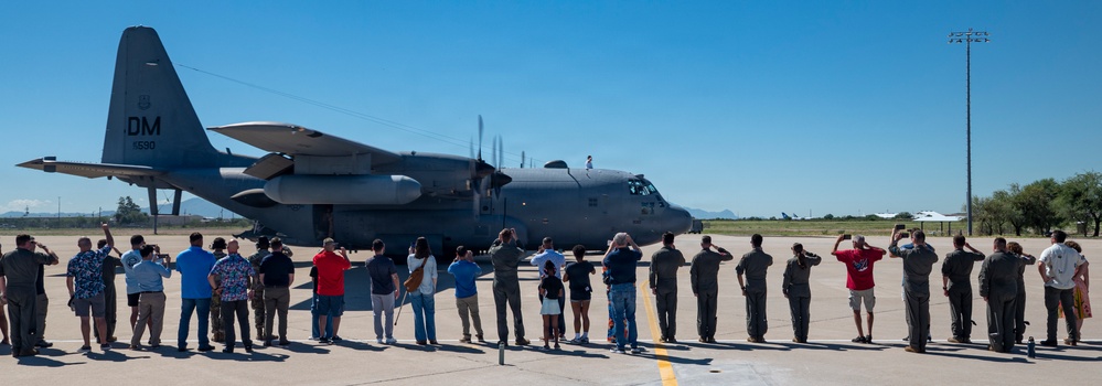 A group of people stands on an airfield tarmac watching and photographing a military turboprop aircraft, identifiable as a C-130 Hercules with tail markings 'DM' and serial number visible. The scene appears to be a formal or ceremonial event, possibly a send-off, homecoming, or retirement ceremony at a military air base. The setting features clear blue skies and desert vegetation consistent with a southwestern U.S. installation.