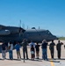 A group of people stands on an airfield tarmac watching and photographing a military turboprop aircraft, identifiable as a C-130 Hercules with tail markings 'DM' and serial number visible. The scene appears to be a formal or ceremonial event, possibly a send-off, homecoming, or retirement ceremony at a military air base. The setting features clear blue skies and desert vegetation consistent with a southwestern U.S. installation.