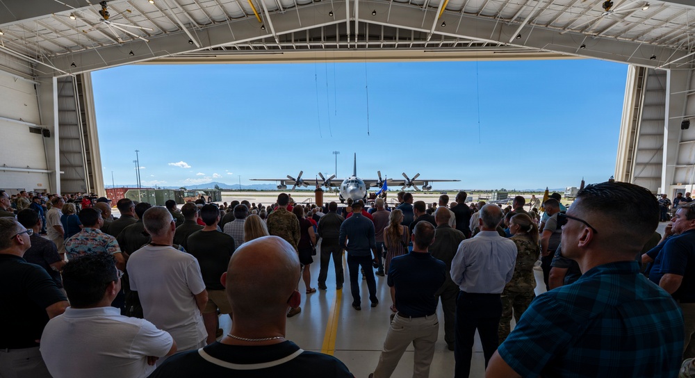 A large crowd of civilians and military personnel gather inside an aircraft hangar, facing outward toward the tarmac where a C-130 Hercules turboprop military transport aircraft is parked. The event appears to be a formal military ceremony or arrival/departure event, likely at a U.S. Air Force or Air National Guard base. Mountains are faintly visible in the background under a clear blue sky.