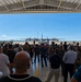 A large crowd of civilians and military personnel gather inside an aircraft hangar, facing outward toward the tarmac where a C-130 Hercules turboprop military transport aircraft is parked. The event appears to be a formal military ceremony or arrival/departure event, likely at a U.S. Air Force or Air National Guard base. Mountains are faintly visible in the background under a clear blue sky.