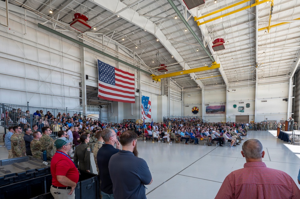 A large military ceremony or formation is taking place inside an aircraft hangar, with hundreds of attendees including uniformed military personnel and civilians. A large American flag is prominently displayed on the back wall alongside what appears to be a unit or organizational flag. A speaker at a podium on the right side addresses the assembled crowd.