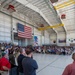 A large military ceremony or formation is taking place inside an aircraft hangar, with hundreds of attendees including uniformed military personnel and civilians. A large American flag is prominently displayed on the back wall alongside what appears to be a unit or organizational flag. A speaker at a podium on the right side addresses the assembled crowd.