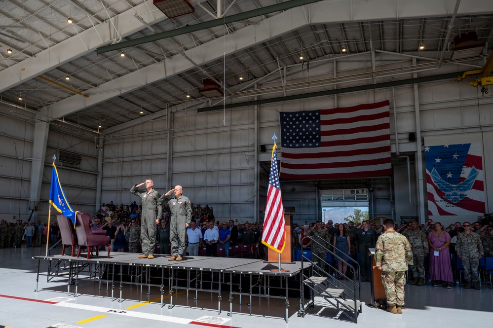 A formal military ceremony is taking place inside a large aircraft hangar. Two officers in flight suits are standing on a raised stage saluting, while a large crowd of military personnel and civilians observe. An American flag is prominently displayed both hanging on the hangar wall and on a flagpole near the podium, suggesting this is likely a change of command or similar official ceremony.